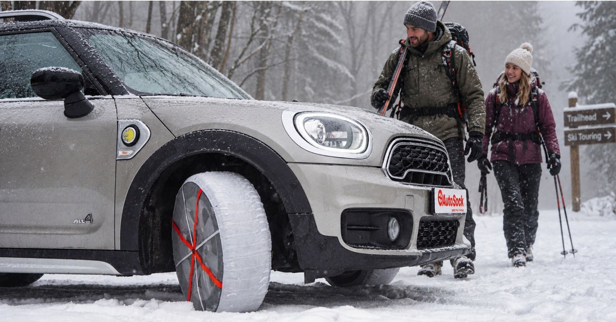 Une Mini Cooper dans une forêt enneigée, équipée de chaussettes à neige. À l’arrière-plan, une femme et un homme reviennent d’une sortie au ski et marchent vers la voiture.